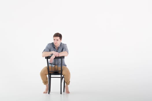 Man dressed in a shirt and brown trousers sits on a wooden vintage chair on a white background