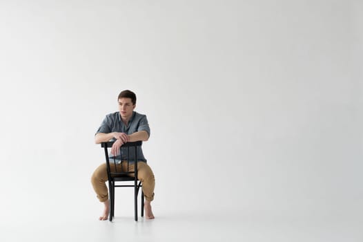 Man dressed in a shirt and brown trousers sits on a wooden vintage chair on a white background
