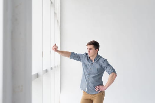 Young handsome man of strong athletic build, dressed in a shirt and brown trousers, Posing on white studio background, rested hand near window in full wall
