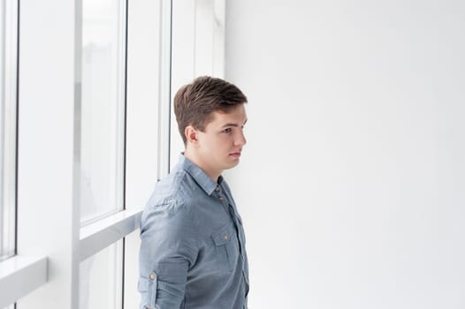 Young handsome man of strong athletic build, dressed in a shirt and brown trousers, Posing on white studio background, near window in full wall, hugging her back