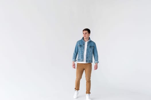 Young handsome man of strong Athlete's body, dressed in Blue jeans jacket in style of old school and white t-shirt with brown trousers, Posing on white studio background