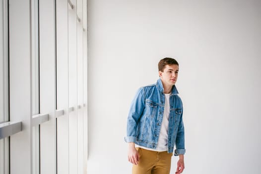 Young handsome man of strong Athlete's body, dressed in Blue jeans jacket in style of old school and white t-shirt with brown trousers, Posing on white studio background, Near a large light window