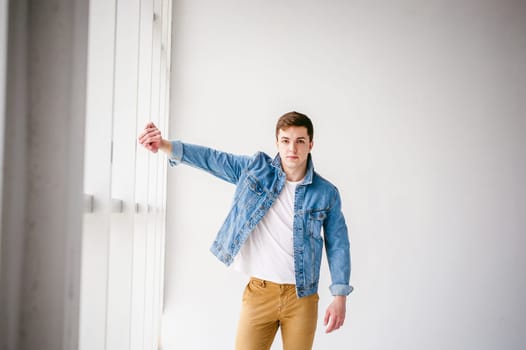 Young handsome man of strong Athlete's body, dressed in Blue jeans jacket in style of old school and white t-shirt with brown trousers, Posing on white studio background, Near a large light window