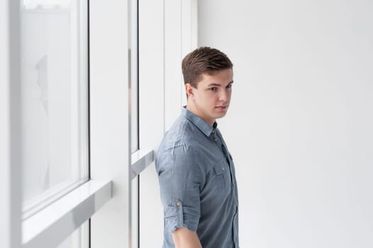 Young handsome man of strong athletic build, dressed in a shirt and brown trousers, Posing on white studio background, near window in full wall, hugging her back
