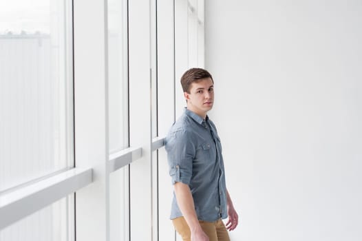 Young handsome man of strong athletic build, dressed in a shirt and brown trousers, Posing on white studio background, near window in full wall, hugging her back