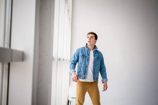 Young handsome man of strong Athlete's body, dressed in Blue jeans jacket in style of old school and white t-shirt with brown trousers, Posing on white studio background, Near a large light window