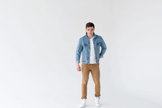 Young handsome man of strong Athlete's body, dressed in Blue jeans jacket in style of old school and white t-shirt with brown trousers, Posing on white studio background