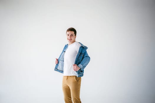 Young handsome man of strong Athlete's body, dressed in Blue jeans jacket in style of old school and white t-shirt with brown trousers, Posing on white studio background