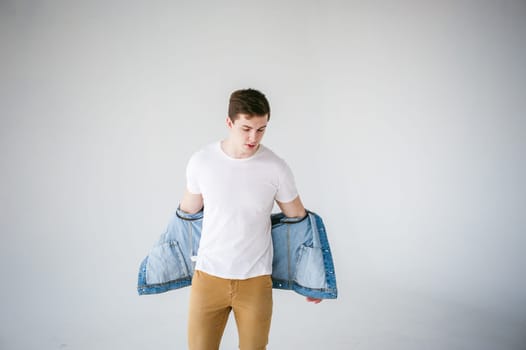 Young handsome man of strong Athlete's body, dressed in Blue jeans jacket in style of old school and white t-shirt with brown trousers, Posing on white studio background