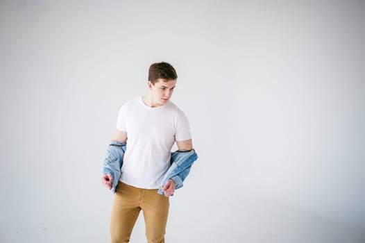 Young handsome man of strong Athlete's body, dressed in Blue jeans jacket in style of old school and white t-shirt with brown trousers, Posing on white studio background