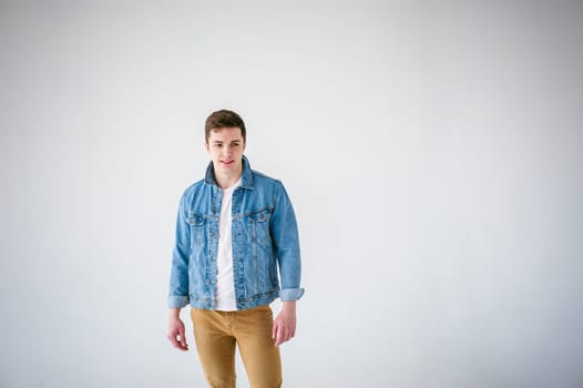 Young handsome man of strong Athlete's body, dressed in Blue jeans jacket in style of old school and white t-shirt with brown trousers, Posing on white studio background