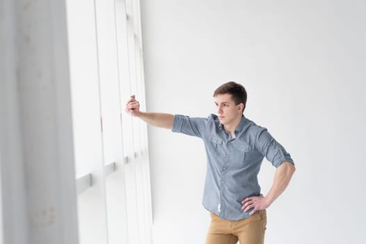 Young handsome man of strong athletic build, dressed in a shirt and brown trousers, Posing on white studio background, rested hand near window in full wall