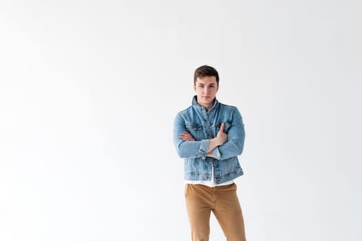 Young handsome man of strong Athlete's body, dressed in Blue jeans jacket in style of old school and white t-shirt with brown trousers, Posing on white studio background, With his arms crossed