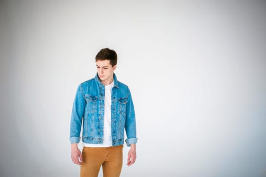 Young handsome man of strong Athlete's body, dressed in Blue jeans jacket in style of old school and white t-shirt with brown trousers, Posing on white studio background