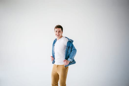 Young handsome man of strong Athlete's body, dressed in Blue jeans jacket in style of old school and white t-shirt with brown trousers, Posing on white studio background