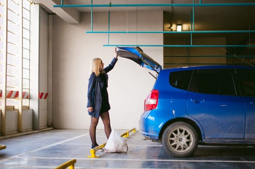 blonde woman in raincoat and pantyhose stacks purchases from grocery store in trunk of the car blue color in covered parking of shopping center
