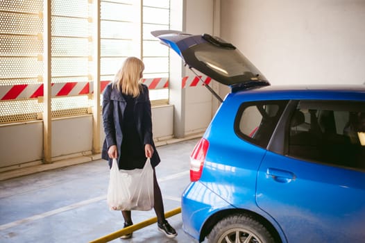 blonde woman in raincoat and pantyhose stacks purchases from grocery store in trunk of the car blue color in covered parking of shopping center