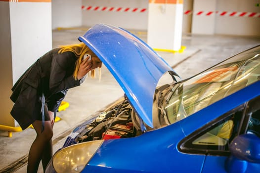woman near car's hood. young blonde in covered parking of shopping center, stands near car with raised engine compartment hood, solves problem with malfunction by phone call to service