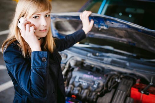 Portrait of blonde woman in black cloak calling phone in car service for help, lifting hood of car and looking at engine, malfunction, diagnosis, service