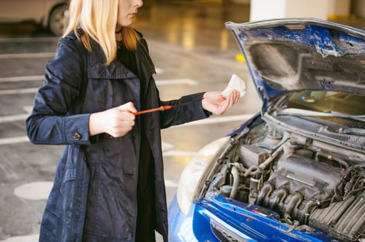 woman near car's hood. young blonde in covered parking of shopping center, stands near car with raised engine compartment hood, checks engine oil level in engine, inspects feeler gauge