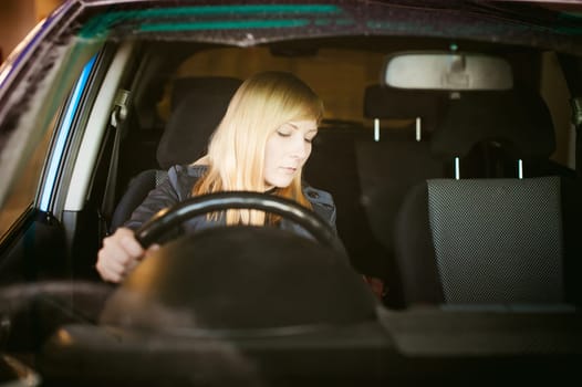 Portrait blonde woman sitting behind wheel in driver's seat of blue car. Hands on handlebars, driving problems of female