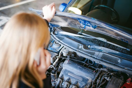 woman near car's hood. young blonde in covered parking of shopping center, stands near car with raised engine compartment hood, solves problem with malfunction by phone call to service