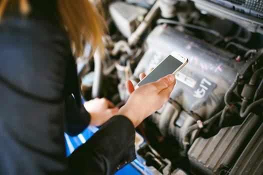 Portrait of blonde woman in black cloak calling phone in car service for help, lifting hood of car and looking at engine, malfunction, diagnosis, service