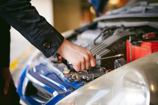woman near car's hood. young blonde in covered parking of shopping center, stands near car with engine compartment bonnet raised, solves problem with engine malfunction, using carabiner wrench tool