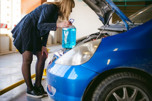 woman near car's hood. young blonde in covered parking of shopping center, stands near car with raised engine compartment hood, pours liquid to wash glass in tank, holding cone in hands