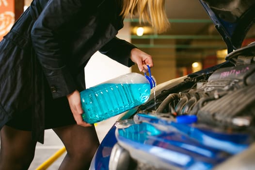 woman near car's hood. young blonde in covered parking of shopping center, stands near car with raised engine compartment hood, pours liquid to wash glass in tank, holding cone in hands