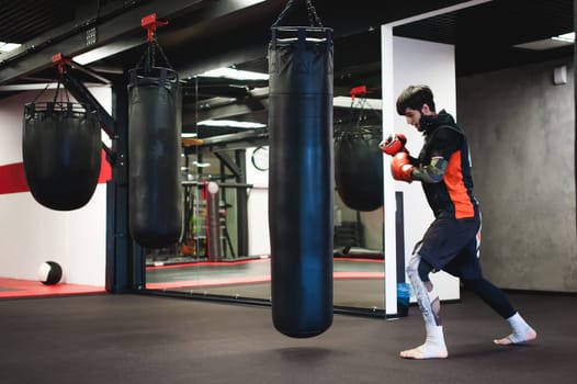 Professional Muay Thai fighter. Male boxer in training sportswear, an exercise in sports hall, Training with a punching bag, working out blows
