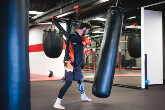 Professional Muay Thai fighter. Male boxer in training sportswear, an exercise in sports hall, Training with a punching bag, working out blows