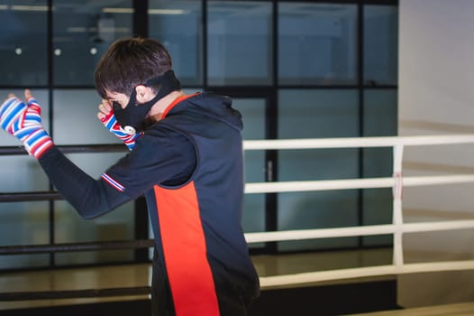 Professional Muay Thai fighter. Male boxer in training sportswear, an exercise in sports hall, fight with a shadow in ring, Strike testing in sports mask
