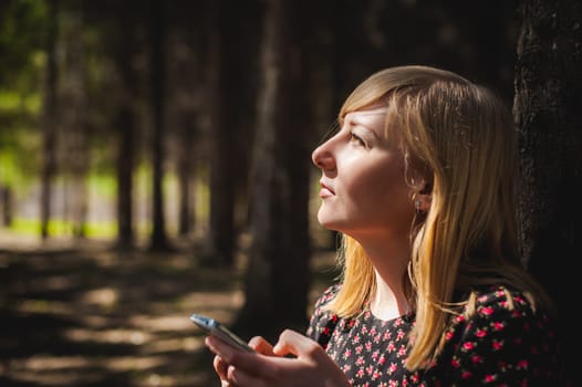 blonde woman dressed summer dress, black pantyhose and sneakers with white sole, resting nature enjoying warm sunny day. Sits under tree alone, holding in hand cell phone, Emotion online communication