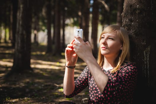 blonde woman dressed summer dress, black pantyhose and sneakers with white sole, resting nature enjoying warm sunny day. Sits under tree alone, holding in hand cell phone, Emotion online communication