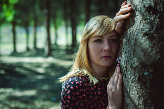 Close-up portrait of blonde woman Hands at head dressed in summer dress. Girl in woods against background of coniferous trees in open air, natural beauty, sunlight shadows on face from branches