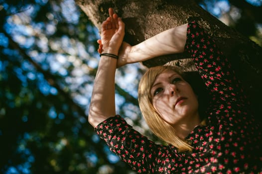 Close-up portrait of blonde woman Hands at head dressed in summer dress. Girl in woods against background of coniferous trees in open air, natural beauty, sunlight shadows on face from branches