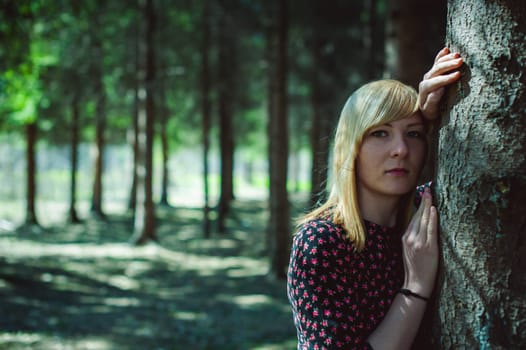 Close-up portrait of blonde woman Hands at head dressed in summer dress. Girl in woods against background of coniferous trees in open air, natural beauty, sunlight shadows on face from branches