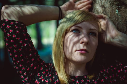 Close-up portrait of blonde woman Hands at head dressed in summer dress. Girl in woods against background of coniferous trees in open air, natural beauty, sunlight shadows on face from branches