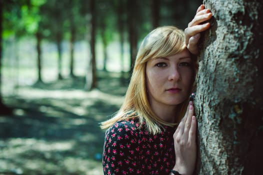 Close-up portrait of blonde woman Hands at head dressed in summer dress. Girl in woods against background of coniferous trees in open air, natural beauty, sunlight shadows on face from branches