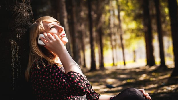 Walk girl coniferous forest park. blonde woman dressed summer dress, black pantyhose and sneakers with white sole, resting nature enjoying warm sunny day. Sits under tree alone, Speaks cell phone