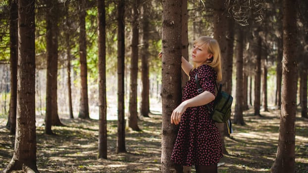 Walk girl coniferous forest park. blond woman with rucksack dressed summer dress, black pantyhose and sneakers with white sole, resting nature enjoying warm sunny afternoon. Stand near tree, hugging