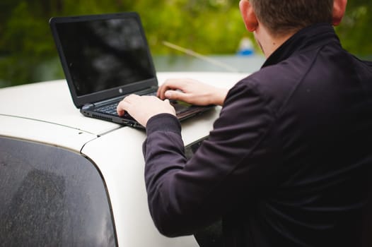 man with a laptop in parking lot in yard near car is doing manipulations with cyber system, concept
