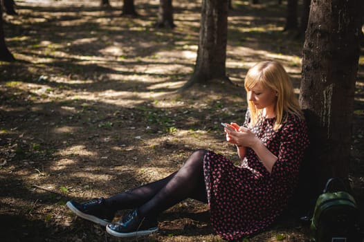 blonde woman dressed summer dress, black pantyhose and sneakers with white sole, resting nature enjoying warm sunny day. Sits under tree alone, holding in hand cell phone, Emotion online communication