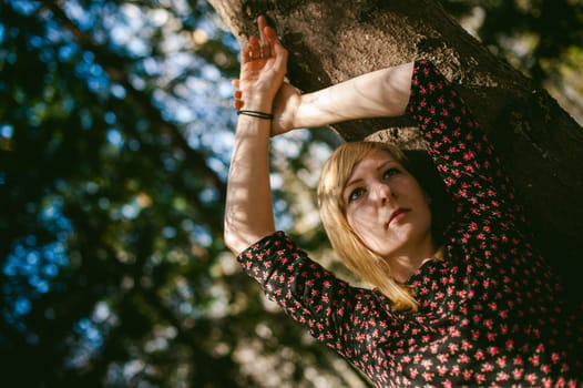 Close-up portrait of blonde woman Hands at head dressed in summer dress. Girl in woods against background of coniferous trees in open air, natural beauty, sunlight shadows on face from branches