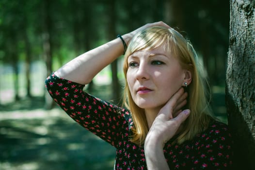 Close-up portrait of blonde woman Hands at head dressed in summer dress. Girl in woods against background of coniferous trees in open air, natural beauty, sunlight shadows on face from branches