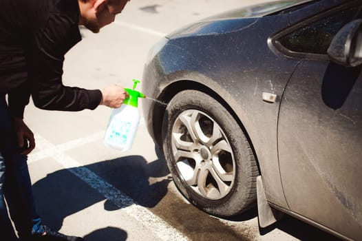 young bearded man washes his car's wheel rims, spraying water from spray, in street parking