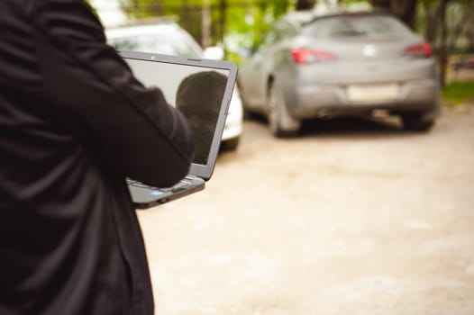 man with a laptop in parking lot in yard near car is doing manipulations with cyber system, concept
