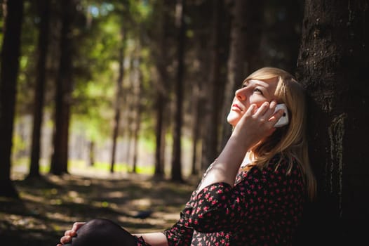Walk girl coniferous forest park. blonde woman dressed summer dress, black pantyhose and sneakers with white sole, resting nature enjoying warm sunny day. Sits under tree alone, Speaks cell phone
