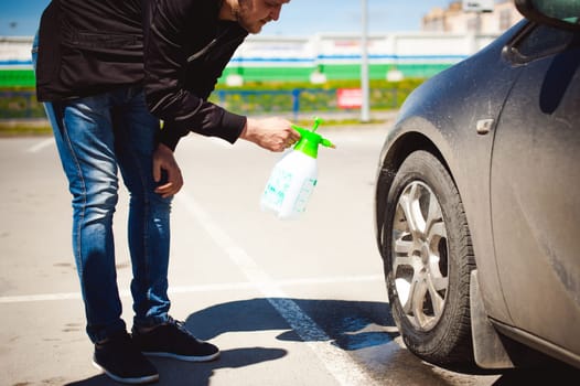 young bearded man washes his car's wheel rims, spraying water from spray, in street parking
